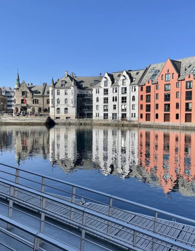 Historic buildings lining the harbour in Ålesund, Norway, reflected in calm water on a clear day.